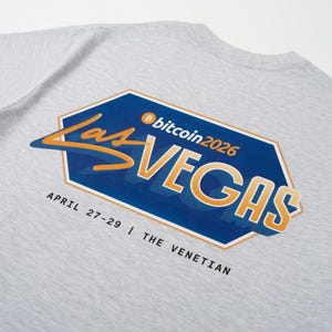 Könnte beinhalten: Ein hellgraues T-Shirt mit einer blauen sechseckigen Grafik und dem orange-weißen Schriftzug Bitcoin 2026 Las Vegas. Darunter steht in schwarzer Schrift April 27-29 | The Venetian. Das Design ist mittig auf der Rückseite des T-Shirts platziert.