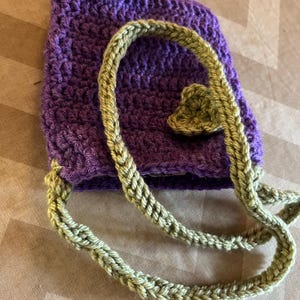 May include: A handmade crochet bag in a deep purple color. The bag features a braided olive green strap and a decorative leaf accent. The crochet work is visible, showcasing the texture and craftsmanship of the bag.