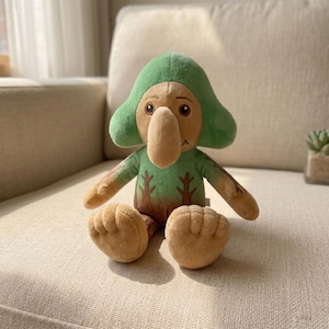 Puede incluir: Una muñeca de peluche con un diseño caprichoso. Presenta un sombrero verde, una larga nariz beige y una camisa verde con diseños de árboles. La muñeca tiene manos y pies beige y está sentada en un sofá beige.