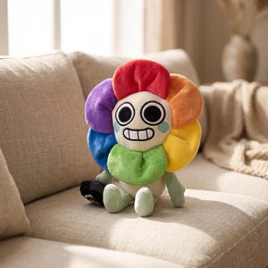 Puede incluir: Un peluche de flor con una cara sonriente y pétalos coloridos en rojo, naranja, amarillo, verde, azul y morado. El juguete tiene un tallo verde y pies verde claro, y está sobre un sofá beige.