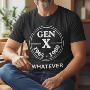 Op de afbeelding: Een zwart t-shirt met korte mouwen voorzien van een witte cirkelvormige afbeelding met de tekst GEN X, de jaren 1965-1980 en het woord WHATEVER in een strak, schreefloos lettertype eronder.