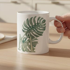 May include: A white ceramic mug featuring a large, light green letter M overlaid with a dark green monstera leaf illustration. The mug has a curved handle and a smooth finish, displaying a botanical design on its exterior surface.