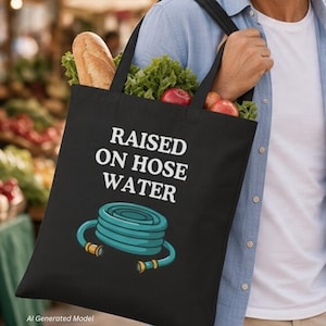 Op de afbeelding: Een zwarte draagtas met de witte tekst RAISED ON HOSE WATER boven een afbeelding van een opgerolde blauwgroene tuinslang met goudkleurige koppelingen. De tas is gevuld met verse producten, waaronder een brood en groene sla.