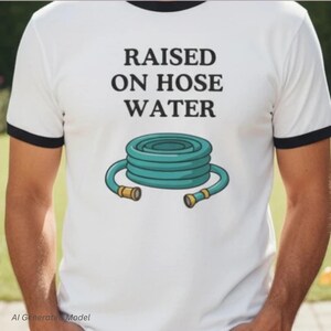 Op de afbeelding: Een wit ringer-t-shirt met zwarte boorden aan de mouwen en kraag. De voorzijde bevat de zwarte tekst RAISED ON HOSE WATER boven een grafische illustratie van een opgerolde blauwgroene tuinslang met goudkleurige metalen koppelingen.