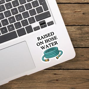 Op de afbeelding: Een witte sticker met een opgerolde turquoise tuinslang met goudkleurige metalen koppelingen aan beide uiteinden. Boven de slang staat de tekst RAISED ON HOSE WATER in dikke, zwarte hoofdletters gedrukt op de sticker.