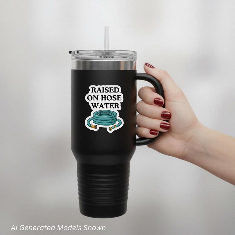 Op de afbeelding: Een zwarte roestvrijstalen beker met een handvat en een doorzichtig deksel met een rietje. De beker heeft een witte sticker met de tekst RAISED ON HOSE WATER en een grafische illustratie van een opgerolde blauwe tuinslang.
