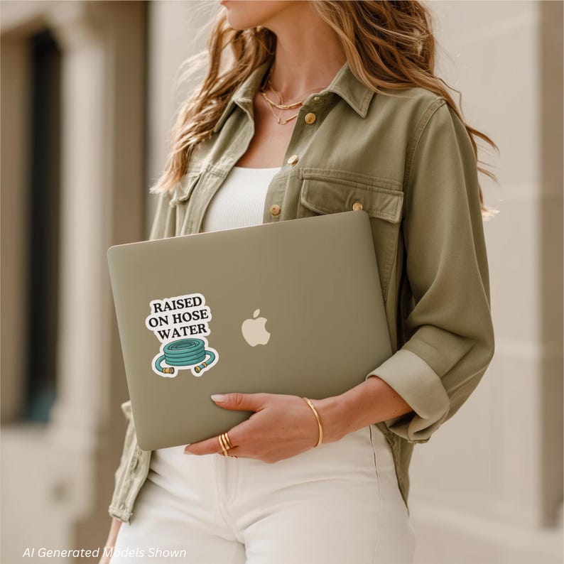 Op de afbeelding: Een mat olijfgroene laptop met een witte, uitgesneden sticker met een illustratie van een blauwgroene tuinslang en de zwarte tekst "RAISED ON HOSE WATER" gecentreerd boven de slangafbeelding, aangebracht op het midden van de laptop.