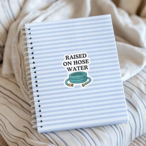 Op de afbeelding: Een spiraalgebonden notitieboek met een lichtblauw en wit horizontaal gestreepte kaft heeft een centrale sticker. De sticker toont een opgerolde blauwgroene tuinslang met gele uiteinden en de zwarte tekst RAISED ON HOSE WATER.