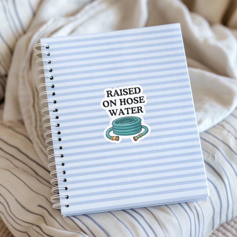 Op de afbeelding: Een spiraalgebonden notitieboek met een lichtblauw en wit horizontaal gestreepte kaft heeft een centrale sticker. De sticker toont een opgerolde blauwgroene tuinslang met gele uiteinden en de zwarte tekst RAISED ON HOSE WATER.