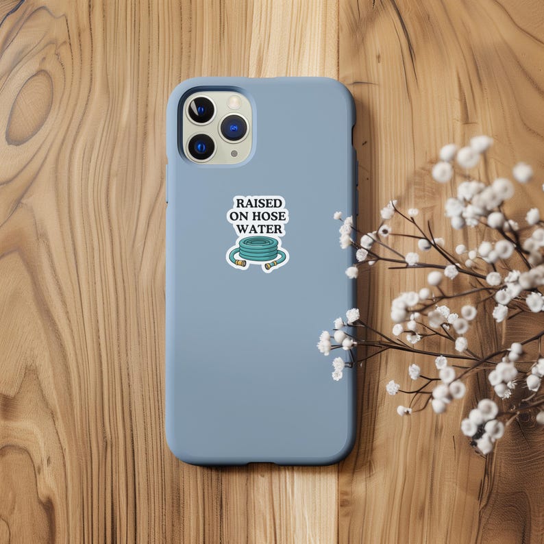Op de afbeelding: Een lichtblauwe smartphonehoes met een sticker met de tekst "RAISED ON HOSE WATER" boven een illustratie van een opgerolde groene tuinslang. De hoes heeft een uitsparing voor een drievoudige camera en een gladde, matte afwerking.