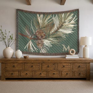 May include: A woven wall tapestry featuring a green background with diagonal stripes, a detailed illustration of a pine branch with two brown pinecones, and a large capital letter M in the lower right corner, finished with fringed edges.