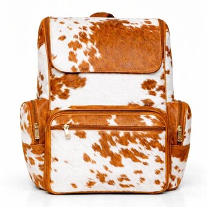 Può includere: Zaino in pelle di vacca con un motivo maculato marrone e bianco. La borsa ha una chiusura a patta, due tasche laterali e una tasca frontale, tutte con cerniere dorate. Il manico superiore e le finiture sono marroni.