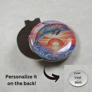 May include: A round, silver-colored golf ball marker with a colorful dolphin design. The dolphin is jumping out of the ocean with a sunset in the background. The text "Personalize it on the back!" is also visible.