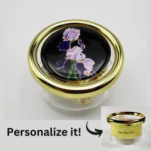 May include: A gold-rimmed glass music box with a black and purple iris flower design on the lid. The lid is transparent and the music box can be personalized with text.