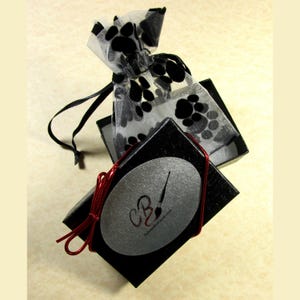 May include: A black gift box with a silver oval sticker that says "CB" and "www.craftycards.com". The box is tied with a red ribbon and has a black and white paw print patterned bag on top.