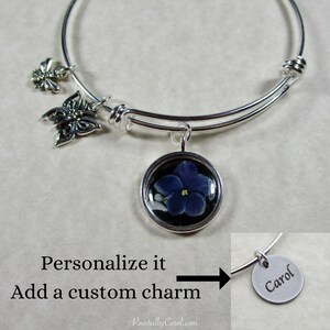 May include: Silver expandable bangle bracelet with a purple flower charm, a butterfly charm, and a bee charm. The bracelet also has a personalized charm with the name "Carol" on it.