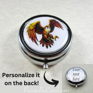 May include: A round, silver-colored pill box with a hinged lid. The lid features a detailed illustration of a hawk with outstretched wings in shades of orange, yellow, and brown against a white background. Text on the image reads "Personalize it on the back!" and "Your text here".