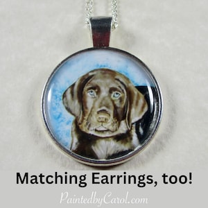 May include: A silver necklace with a round pendant featuring a painted portrait of a chocolate brown Labrador Retriever puppy. The pendant has a blue and white background.