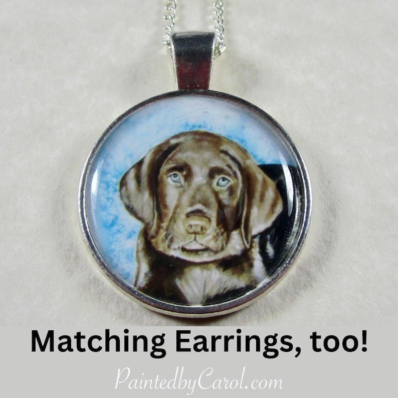 May include: A silver necklace with a round pendant featuring a painted portrait of a chocolate brown Labrador Retriever puppy. The pendant has a blue and white background.