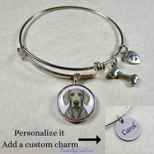 May include: A silver-tone expandable bangle bracelet featuring a circular pendant with a Weimaraner dog illustration, a small silver bone charm, and a heart-shaped paw print charm. An inset shows a round silver charm engraved with the name Carol.