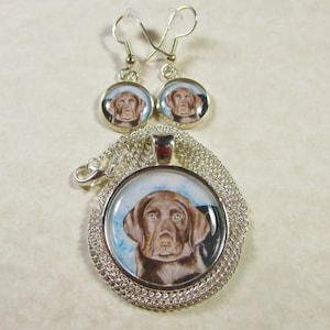 May include: A silver necklace and earrings set featuring a round pendant with a painted image of a chocolate brown Labrador Retriever puppy. The pendant and earrings have a silver chain and a silver hook.