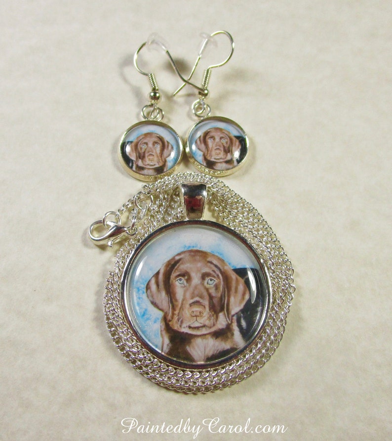 May include: A silver necklace and earrings set featuring a round pendant with a painted image of a chocolate brown Labrador Retriever puppy. The pendant and earrings have a silver chain and a silver hook.