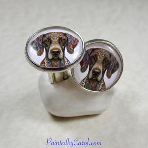May include: A pair of round silver-toned cufflinks featuring a detailed illustration of a brown dog with intricate, colorful, paisley-like patterns on its ears and face. The dog has a white muzzle and dark eyes, set against a plain white background.