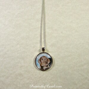 May include: A silver chain necklace with a round pendant featuring a watercolor painting of a brown dog with blue eyes. The pendant is set in a silver frame.