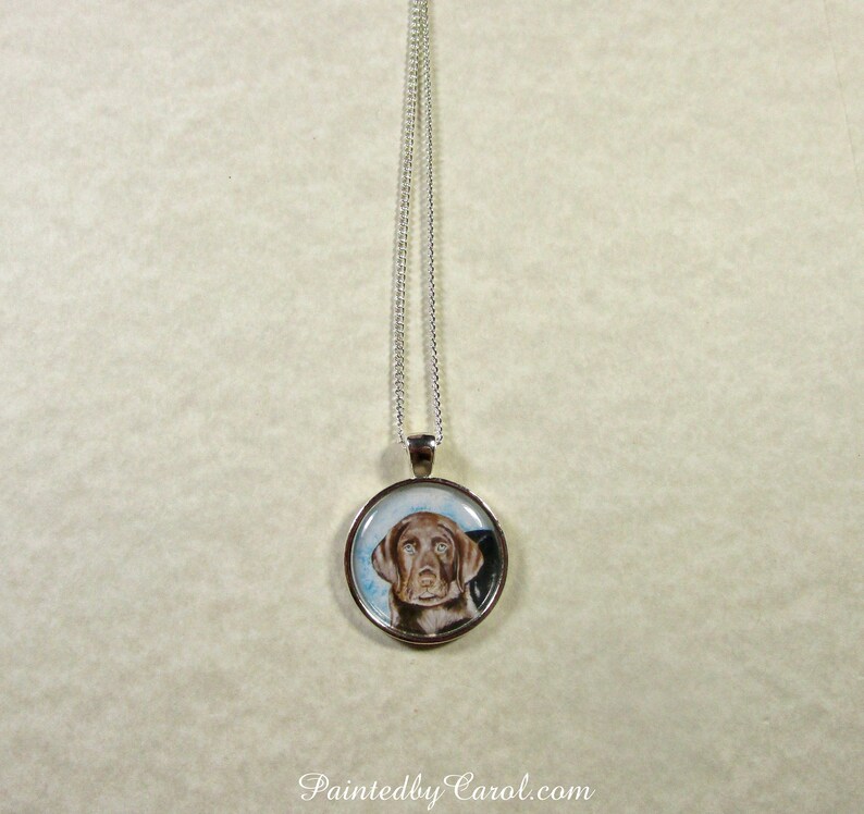 May include: A silver chain necklace with a round pendant featuring a watercolor painting of a brown dog with blue eyes. The pendant is set in a silver frame.