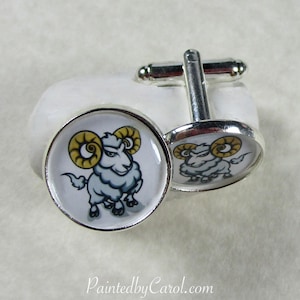 May include: A pair of silver cufflinks with a white background and a cartoon image of a white ram with yellow horns. The cufflinks are on a white surface.