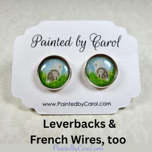 May include: A pair of round silver-toned earrings featuring a painted image of a dog in a grassy field under a blue sky. The earrings are displayed on a white card with the text "Painted by Carol" and "Leverbacks & French Wires, too".