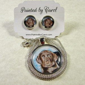 May include: A silver necklace with a round pendant featuring a painted portrait of a chocolate brown Labrador Retriever puppy. The pendant is surrounded by a silver chain. The necklace comes with a pair of matching earrings with the same image.