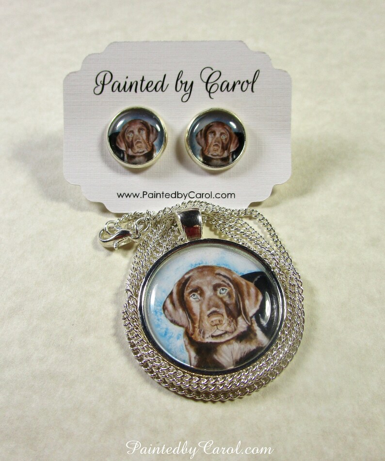 May include: A silver necklace with a round pendant featuring a painted portrait of a chocolate brown Labrador Retriever puppy. The pendant is surrounded by a silver chain. The necklace comes with a pair of matching earrings with the same image.