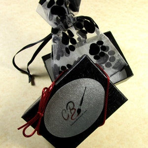 May include: A black box with a silver oval sticker that says "CB" and "PaintedbyCarol.com". The box is tied with a red ribbon and is open to reveal a white mesh bag with black paw prints.