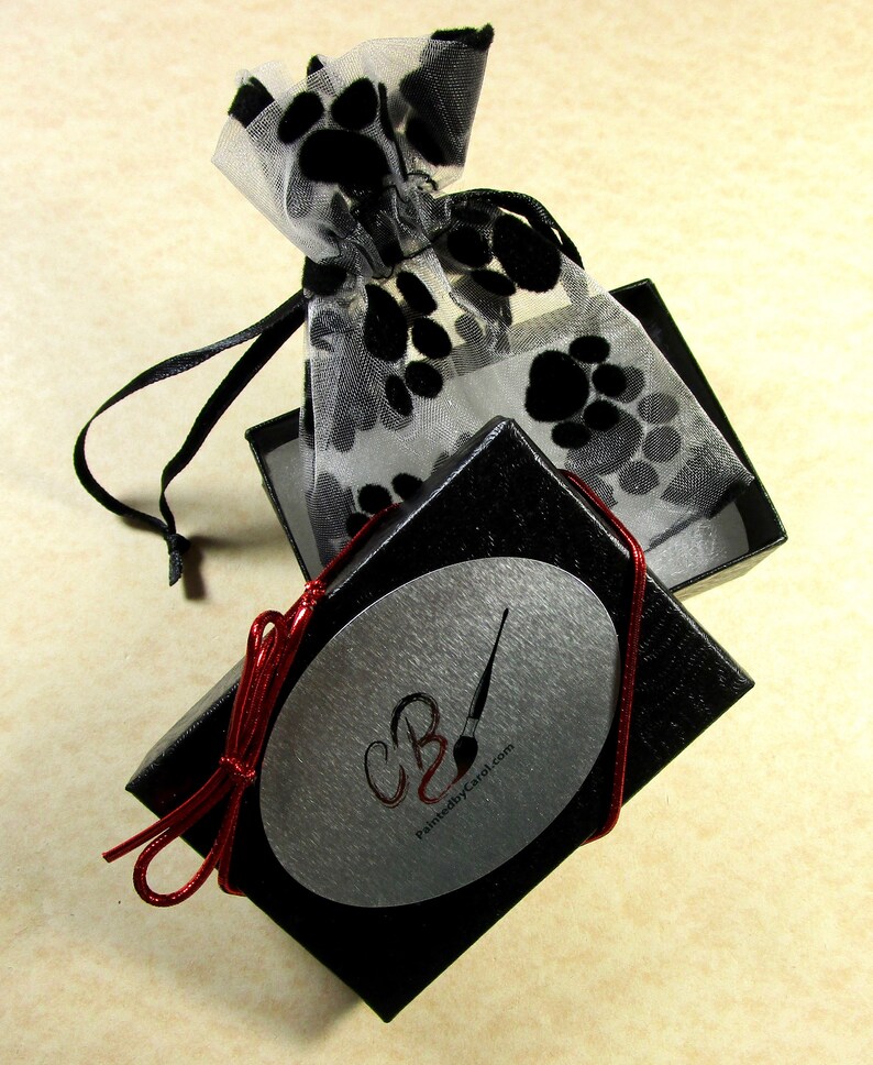 May include: A black box with a silver oval sticker that says "CB" and "PaintedbyCarol.com". The box is tied with a red ribbon and is open to reveal a white mesh bag with black paw prints.