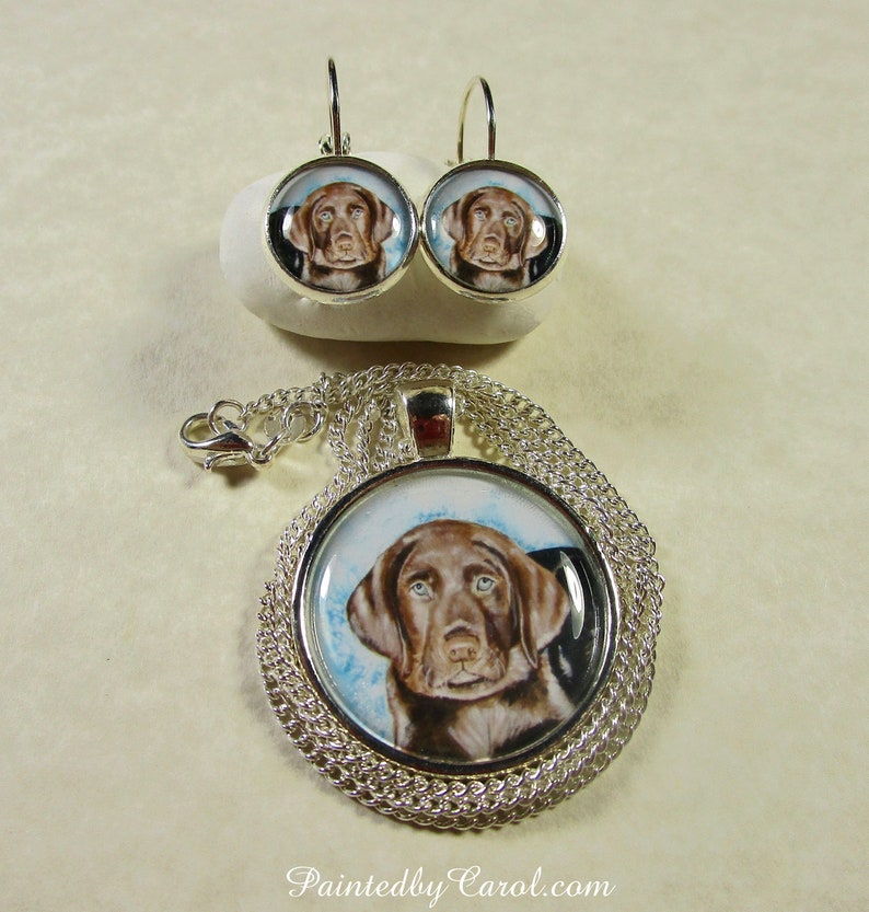 May include: A silver necklace and earrings set featuring a brown and white dog portrait. The necklace has a silver chain and a round pendant with a clear glass front. The earrings are silver hoops with a clear glass front.