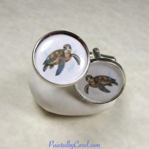 May include: A pair of round silver-toned cufflinks featuring a detailed illustration of a sea turtle with green, brown, and orange markings on a white background. The text PaintedbyCarol.com is printed in blue at the bottom of the image.