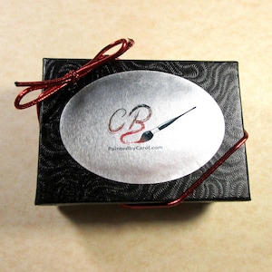 May include: A black gift box with a red ribbon and a silver oval sticker. The sticker has the letters "CB" in red and a paintbrush with a red handle. The text "PaintedbyCarol.com" is printed below the letters.