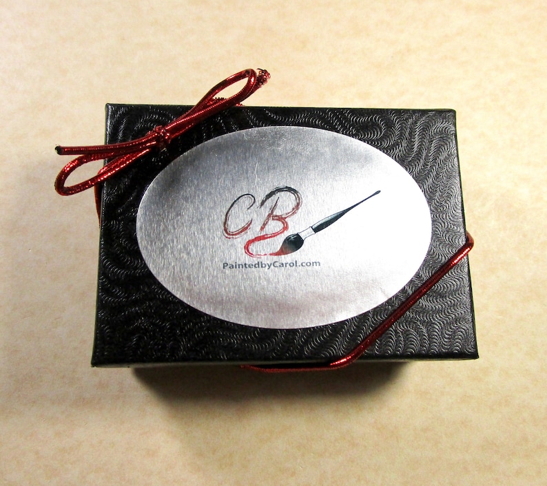 May include: A black gift box with a red ribbon and a silver oval sticker. The sticker has the letters "CB" in red and a paintbrush with a red handle. The text "PaintedbyCarol.com" is printed below the letters.