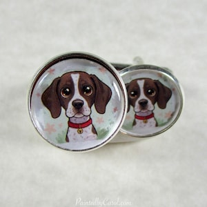 May include: Two silver-toned cufflinks featuring a detailed illustration of a dog's face. The dog has brown and white fur, large brown eyes, and a red collar. The background is a light green with pink floral accents.