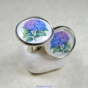 May include: A pair of round silver cufflinks featuring a detailed illustration of a blue and purple hydrangea flower with green leaves on a white background. The metal frames are polished silver, and the design is protected by a clear glass dome.