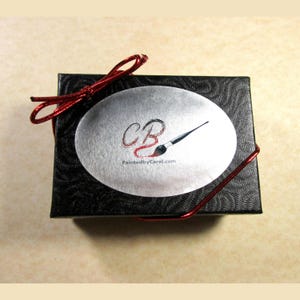 May include: A black gift box with a red ribbon and a silver oval sticker. The sticker has the letters "CB" and a paintbrush with the text "PaintedbyCarol.com".