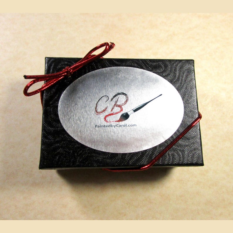 May include: A black gift box with a red ribbon and a silver oval sticker. The sticker has the letters "CB" and a paintbrush with the text "PaintedbyCarol.com".