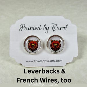 May include: A pair of round stud earrings featuring a detailed illustration of a bear's head in shades of red, brown, and yellow. The earrings have a silver-colored metal border and are displayed on a white card with the text "Painted by Carol".