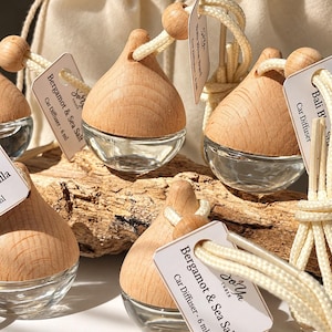 May include: Multiple glass car diffusers with light wood teardrop-shaped caps and hanging cords. Each unit features a small white tag labeled Bergamot & Sea Salt Car Diffuser - 6 ml or Bali Breeze Car Diffuser. The diffusers are clear glass.