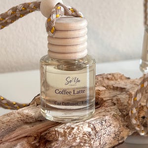 Op de afbeelding: Een doorzichtig glazen 8 ml autodiffuserflesje met een lichthouten dop en een gevlochten beige en goudkleurig koord. Het etiket bevat de tekst So Ya Amber, Coffee Latte en Car Diffuser - 8 ml. Het flesje is gevuld met een lichtgekleurde vloeistof.
