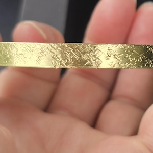 May include: A gold-colored bracelet with a textured surface. The bracelet is held in a hand, showcasing its design. The jewelry piece has a flat, rectangular shape and a detailed, embossed pattern.