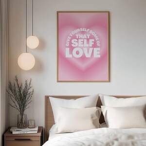 May include: A rectangular wall art print in a gold frame featuring a pink gradient heart background with the white text GIVE YOURSELF SOME OF THAT SELF LOVE centered in the middle of the heart. The design uses a soft, blurred aesthetic.