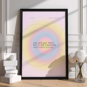 May include: A framed wall poster featuring a soft, gradient aura background in pastel yellow, pink, and blue. The text reads: 2023 poster gallery, and you will get there but in the meantime enjoy where you are. Thin black circular lines overlay the design.