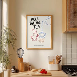 Op de afbeelding: Een ingelijste wandplaat met de zwarte tekst 'HERE FOR THE TEA', een blauwe lijntekening van een theepot die in een kopje schenkt, rode lijntekeningen van gestapelde kopjes met stoom en een kleine groep koekjes in een lichtbruine omlijning.